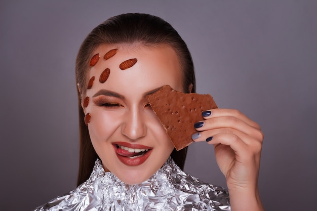 Retrato de uma mulher de cabelos castanho alegre com maquiagem brilhante, segurando a barra de chocolate no rosto, isolada sobre o fundo