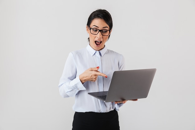 Retrato de uma mulher de negócios adulta usando óculos, segurando um laptop prateado no escritório, isolado sobre uma parede branca