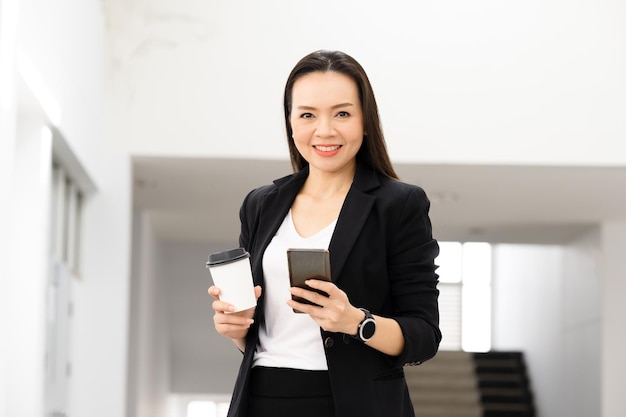 Retrato de uma mulher de negócios asiática de meia-idade segurando um telefone inteligente e um café sorrindo para a câmera