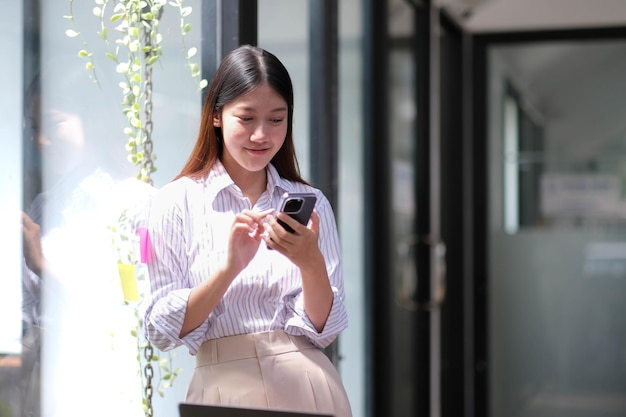 Retrato de uma mulher de negócios asiática elegante e bem-sucedida falando por smartphone e sorrindo alegremente enquanto se posiciona contra a janela no escritório