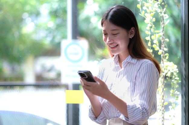 Retrato de uma mulher de negócios asiática elegante e bem-sucedida falando por smartphone e sorrindo alegremente enquanto se posiciona contra a janela no escritório