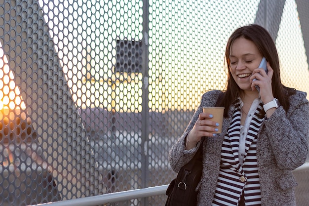 Retrato de uma mulher de negócios falando ao telefone na rua com um café na mão ao pôr do sol