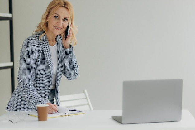 Retrato de uma mulher de negócios madura sentada em um escritório moderno falando ao telefone e escrevendo algo em um caderno