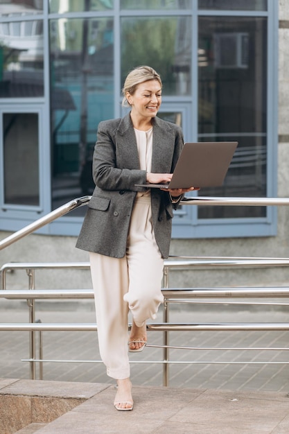 Retrato de uma mulher de negócios maduros sorrindo com emoções e segurando um laptop falando em uma chamada de vídeo e trabalhando no fundo de edifícios urbanos e de escritórios modernos