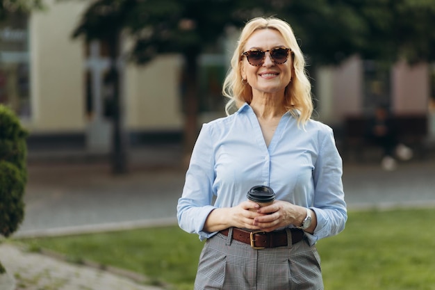 Retrato de uma mulher de negócios sênior segurando um copo de papel Mulher feliz sorridente idosa em seus 50 anos caminha na cidade Conceito de negócio Sucesso no trabalho