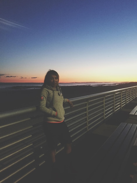 Retrato de uma mulher de pé junto a uma grade contra o céu durante o pôr do sol