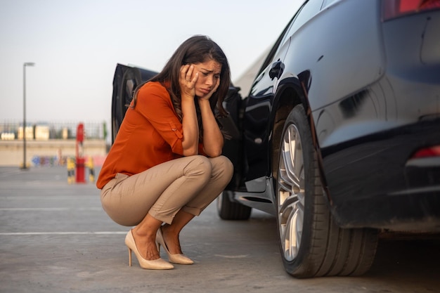 Retrato de uma mulher desesperada agachada ao lado de seu carro com problemas de pneus furados na estrada pneu desinflado