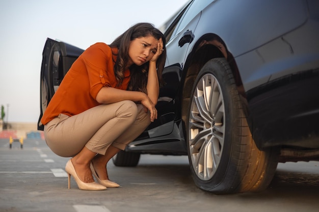 Foto retrato de uma mulher desesperada agachada ao lado de seu carro com problemas de pneus furados na estrada pneu desinflado