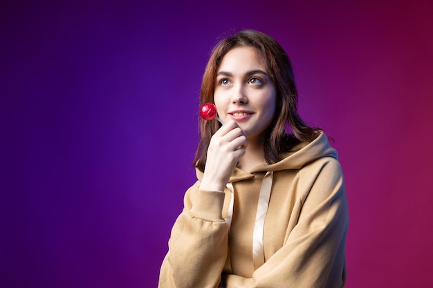 Retrato de uma mulher em um capuz meninas lambendo um pirulito redondo vermelho com bela maquiagem em um fundo roxo na moda no estúdio