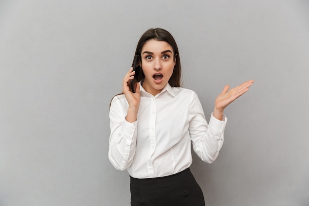 Retrato de uma mulher emocionalmente profissional com longos cabelos castanhos em traje formal enquanto fala em um smartphone, isolado sobre uma parede cinza