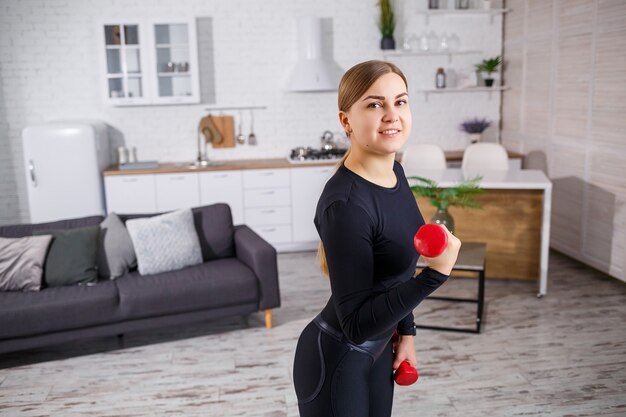 Retrato de uma mulher esbelta vai para a prática de esportes em casa, com a blusa e as leggings com roupas de ginástica. Fitness em casa por um corpo lindo. Exercícios com halteres para o corpo feminino