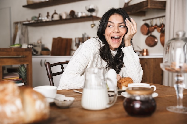 Retrato de uma mulher europeia feliz sentada à mesa e comendo enquanto toma o café da manhã na cozinha de casa