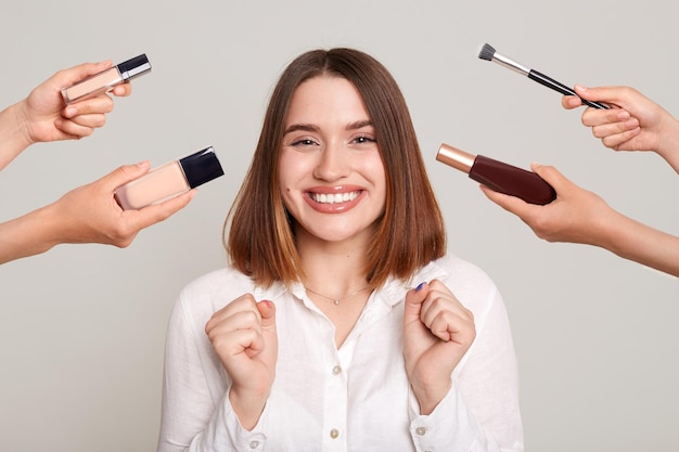 Retrato de uma mulher extremamente feliz com cabelos castanhos escuros em pé com as mãos cerradas, satisfeita com sua maquiagem cercada de mãos com cosméticos