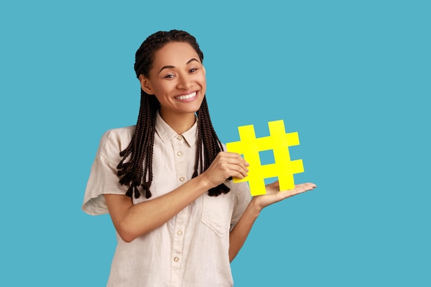 Retrato de uma mulher feliz com dreadlocks pretos mostrando hashtag amarela, comunidade da internet, conteúdo de blog famoso, promoção, vestindo camisa branca. Tiro de estúdio interior isolado sobre fundo azul.