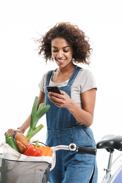 Retrato de uma mulher feliz com uma sacola de compras segurando um celular enquanto andava de bicicleta isolada na parede branca