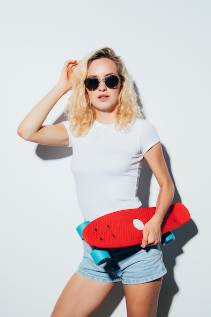 Foto retrato de uma mulher feliz e alegre em óculos de sol, posando com um skate em pé sobre uma parede branca