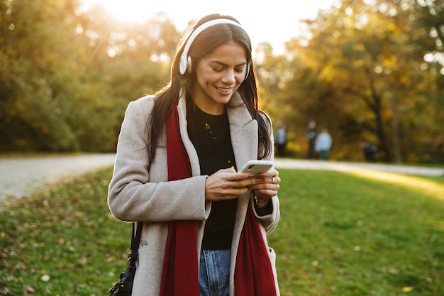 Retrato de uma mulher feliz e simpática vestindo um casaco, ouvindo música com fones de ouvido e usando o celular enquanto caminha no parque outono