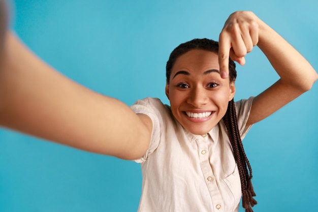 Retrato de uma mulher feliz sorridente com dreadlocks fazendo selfie, apontando o dedo para baixo, pedindo para se inscrever, foto do ponto de vista, vestindo camisa branca. Tiro de estúdio interior isolado sobre fundo azul.