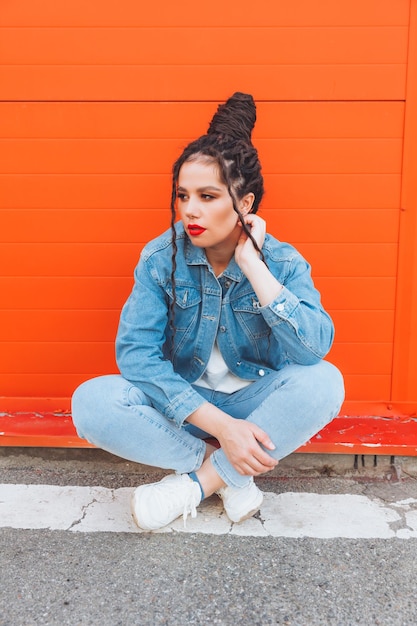 Retrato de uma mulher glamourosa com dreadlocks em um terno jeans e com lábios vermelhos contra uma parede laranja