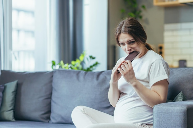 Retrato de uma mulher grávida comendo uma barra de deliciosos doces de chocolate Sentado no sofá em casa com roupas de casa olhando para a câmera sorrindo Satisfeito adora doces