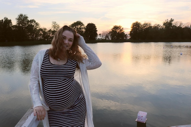 Foto retrato de uma mulher grávida de pé ao lado do lago contra o céu durante o pôr-do-sol