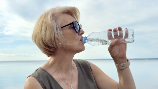Retrato de uma mulher idosa e bonita bebendo água de uma garrafa de plástico no dia de verão em