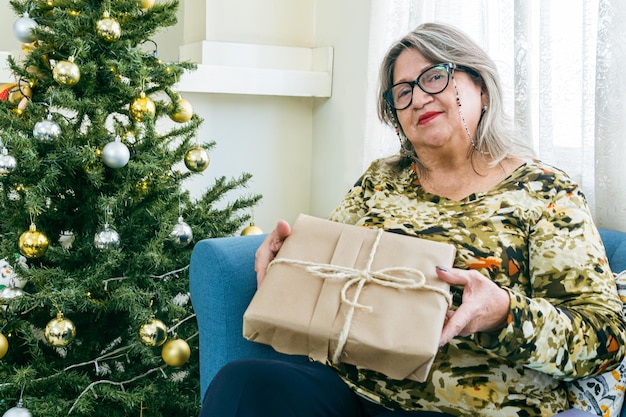 Foto retrato de uma mulher idosa hispânica olhando para a câmera com um presente ao lado da árvore de natal