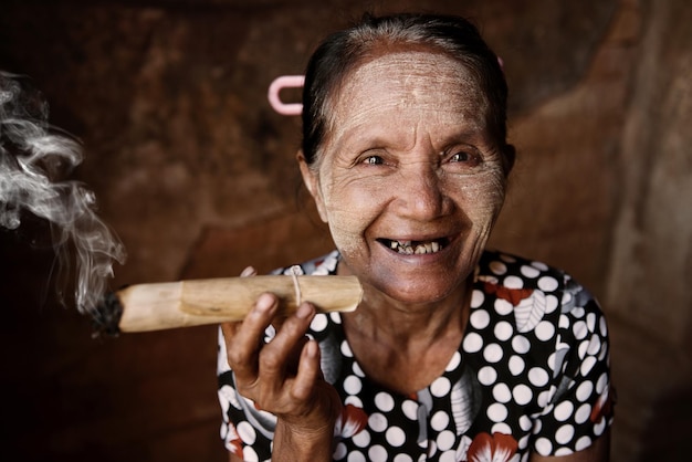 Foto retrato de uma mulher idosa sorridente com tinta facial de thanaka fumando charuto