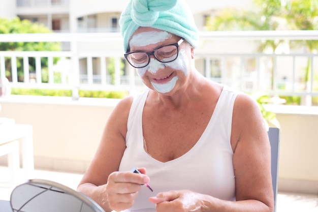 Retrato de uma mulher idosa sorridente de cabelos grisalhos com máscara antienvelhecimento no rosto enrugado aplicando esmalte para unhas