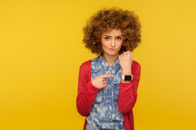 Retrato de uma mulher impaciente chateada com cabelo encaracolado, mostrando o relógio de pulso e parecendo decepcionado com a câmera, lembrando da hora tardia, pedindo para apressar o tiro de estúdio interno isolado no fundo amarelo