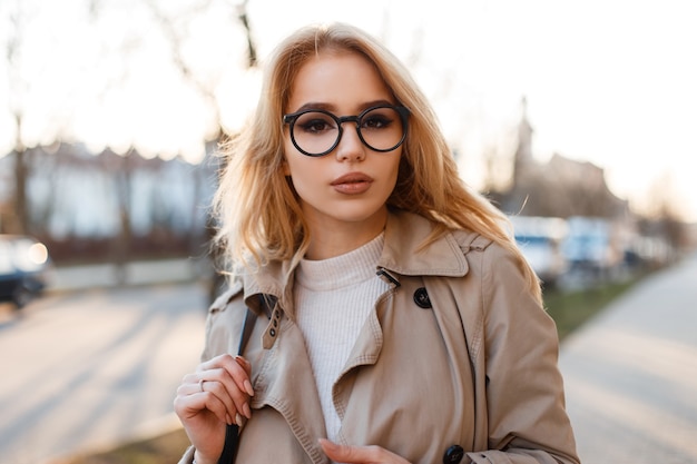 Retrato de uma mulher incrível jovem bonito hippie com belos lábios em copos elegantes com um elegante casaco de primavera em uma camiseta branca na rua durante a tarde de primavera. Garota atraente e elegante.