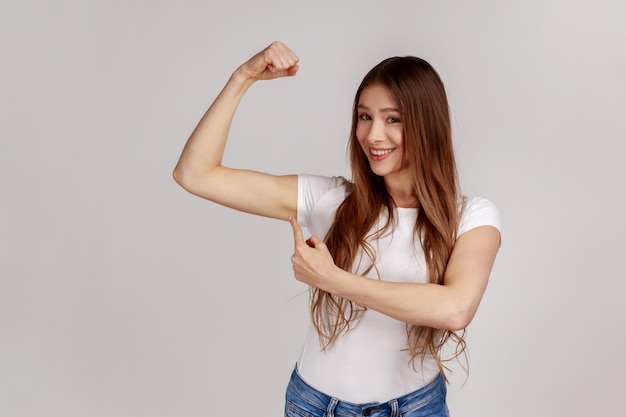 Retrato de uma mulher independente confiante levantando a mão apontando o bíceps, sentindo poder e energia para atingir o objetivo, vestindo camiseta branca. Tiro de estúdio interior isolado em fundo cinza.