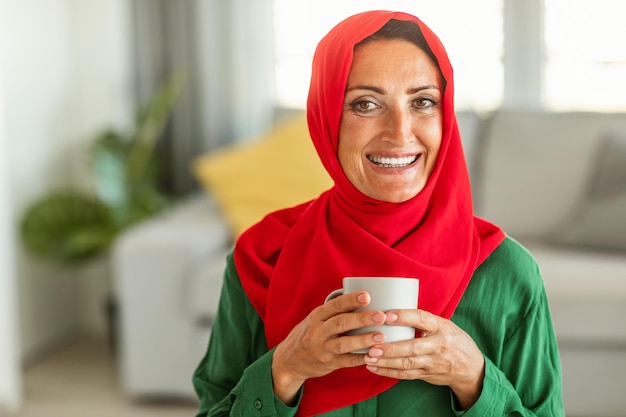 Retrato de uma mulher islâmica de meia-idade feliz em hijab sentada na sala de estar com uma xícara de chá de ervas e sorrindo para a câmera