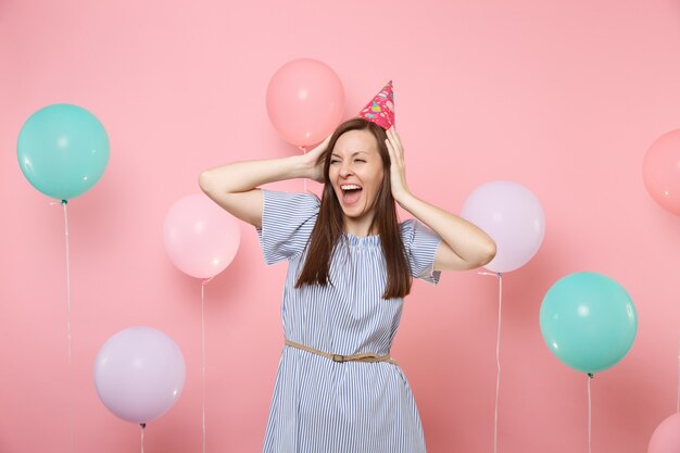 Retrato de uma mulher jovem e atraente alegre com chapéu de aniversário e vestido azul agarrado à cabeça no fundo rosa de tendência brilhante com balões de ar coloridos. Festa de aniversário, emoções sinceras de pessoas.