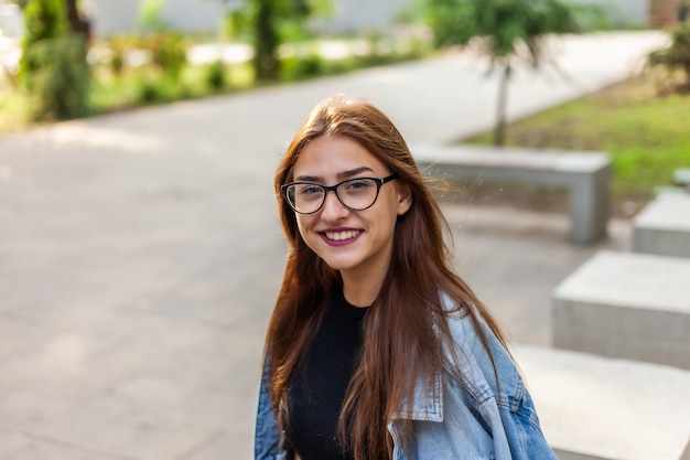 Retrato de uma mulher jovem estudante em uma jaqueta jeans e óculos em um parque ao ar livre