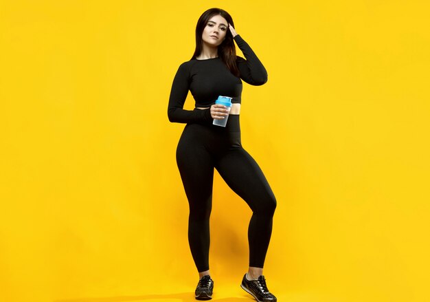 Retrato de uma mulher latina positiva de corpo lindo em um terno preto de esportes com uma garrafa de água posando em amarelo