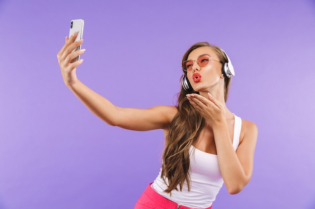 Retrato de uma mulher linda em roupas de verão e óculos de sol, tomando selfie enquanto ouve música usando fones de ouvido sem fio e smartphone