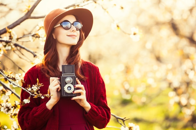 Retrato de uma mulher linda ruiva de suéter vermelho e chapéu com câmera no jardim de macieira flor na primavera no pôr do sol.