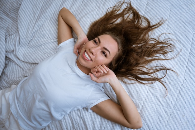 Retrato de uma mulher loira adulta jovem e atraente em uma camiseta branca, deitada na cama com cabelo desgrenhado.