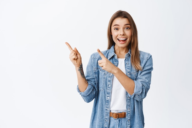 Foto retrato de uma mulher loira animada sorrindo divertido, apontando o dedo para a oferta promocional do canto superior esquerdo, mostrando um anúncio para recomendar uma boa oferta, parede branca