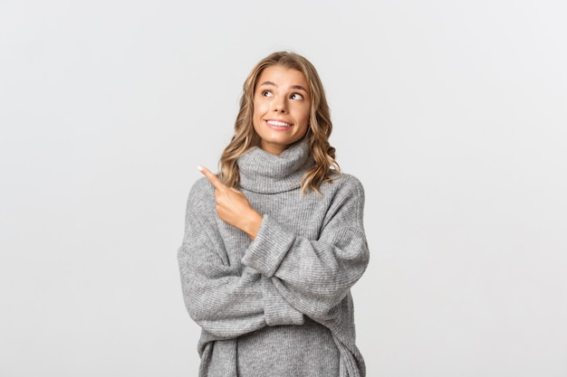 Retrato de uma mulher loira atraente com um suéter cinza, sorrindo, sonhadora e apontando para o canto superior esquerdo