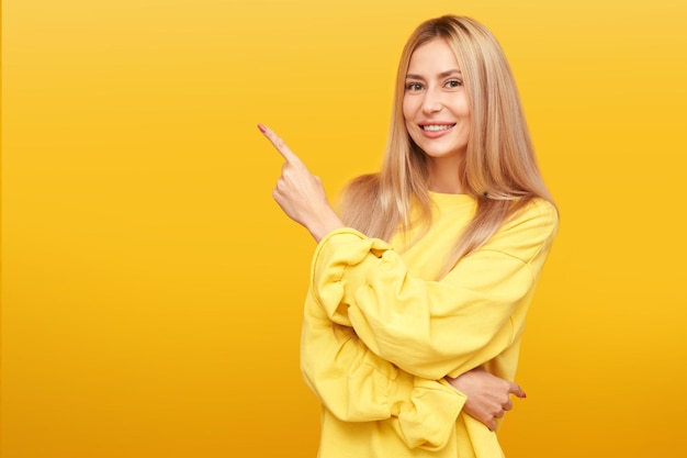 Retrato de uma mulher loira atraente sorrindo e apontando o dedo para o espaço vazio em fundo amarelo isolado