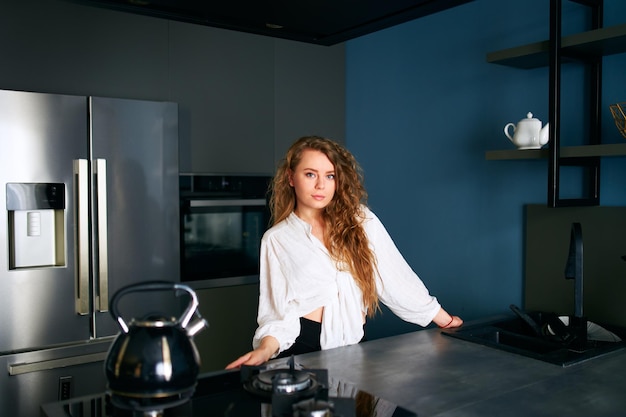 Retrato de uma mulher loira caucasiana de pé e sorrindo em sua cozinha, inclinando-se sobre a mesa jovem