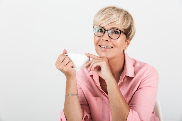 Retrato de uma mulher loira de meia-idade usando óculos, sorrindo e segurando uma xícara de café isolada sobre uma parede branca