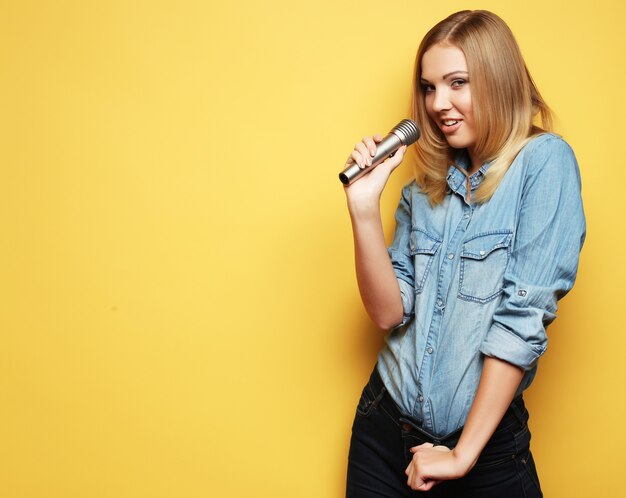 Retrato de uma mulher loira encantadora cantando com microfone sobre o espaço amarelo.