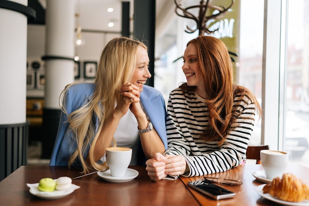 Retrato de uma mulher loira rindo falando com uma amiga ruiva feliz passando tempo juntos enquanto bebem café em um café aconchegante sentada em uma mesa junto à janela Conceito de amizade feminina
