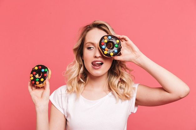 Retrato de uma mulher loira satisfeita vestindo uma camiseta casual sorrindo enquanto segura deliciosos donuts doces isolados sobre uma parede rosa