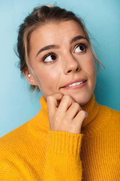 Retrato de uma mulher loira séria com um suéter amarelo, olhando para cima e pensando isolado no azul