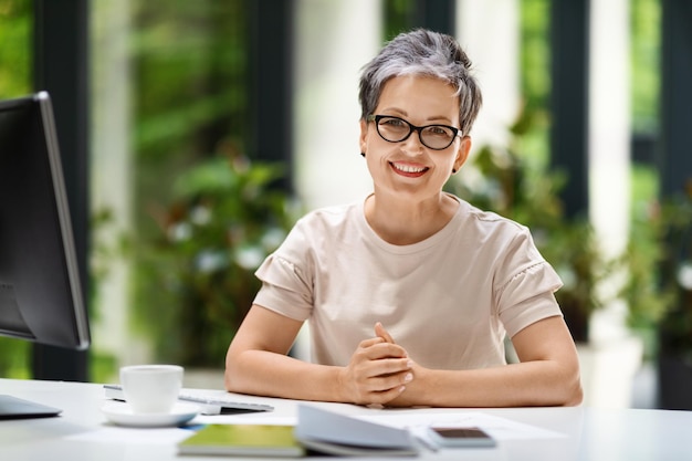 Retrato de uma mulher madura alegre sentada no local de trabalho com um computador