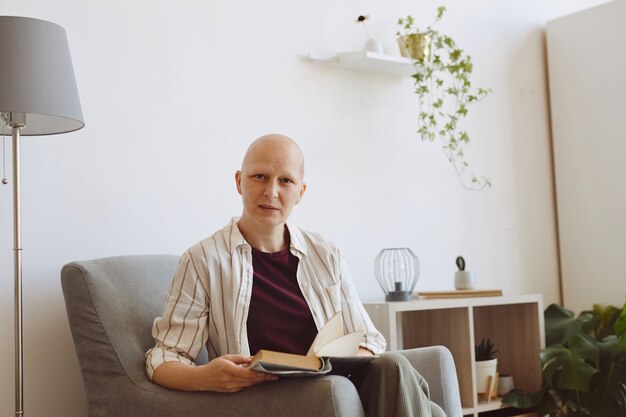 Retrato de uma mulher madura careca olhando para a câmera enquanto lê um livro sentado em uma poltrona confortável em casa. alopecia e conscientização do câncer, copie o espaço